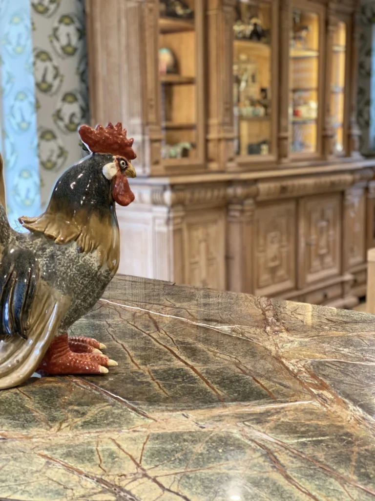 A Ceramic Rooster Figurine Sits On A Polished Marble Countertop. In The Background, There'S A Wooden Cabinet With Various Decorative Items Displayed On Its Shelves. The Scene Is Set In A Room With A Patterned Wallpaper.