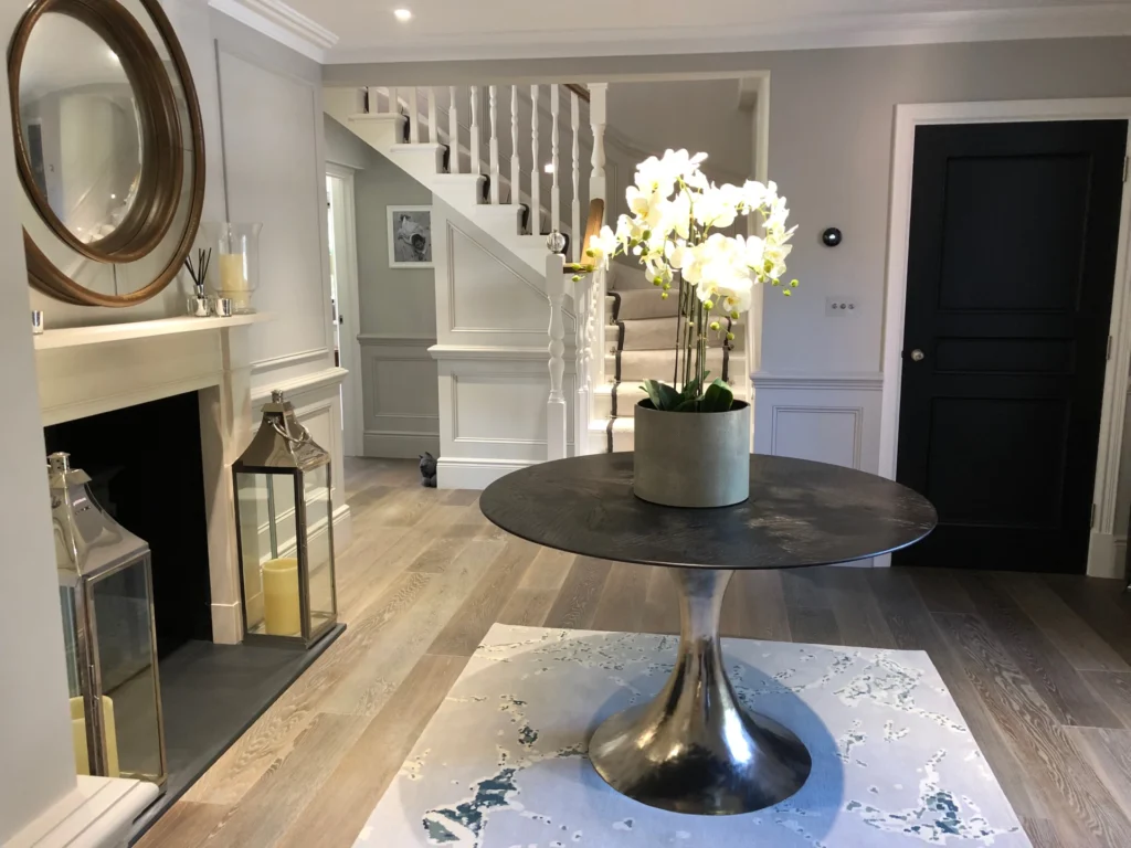 A Modern Entryway Features A Round Metal Table With A Potted Orchid On Top, Placed On A Light Colored Rug. The Background Shows A Staircase With White Railing, A Black Door, Framed Artwork, A Mirror, And A Fireplace With Two Large Lanterns.